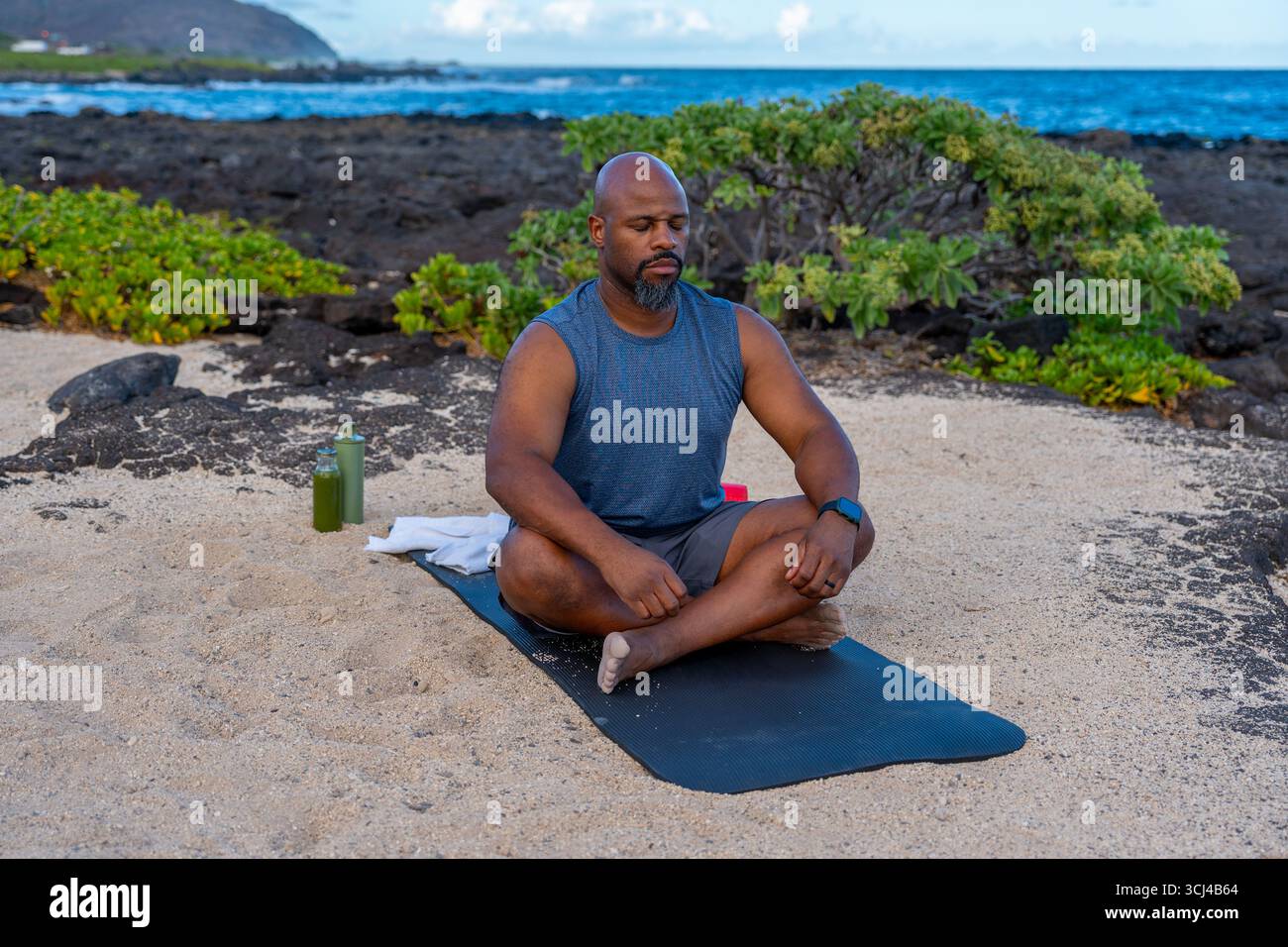Uomo nero che meditava in posa di loto, occhi chiusi a fuoco pacifico. Spiaggia tropicale all'aperto che si avvicina al tramonto con luce soffusa evoca calma, consapevolezza, Foto Stock