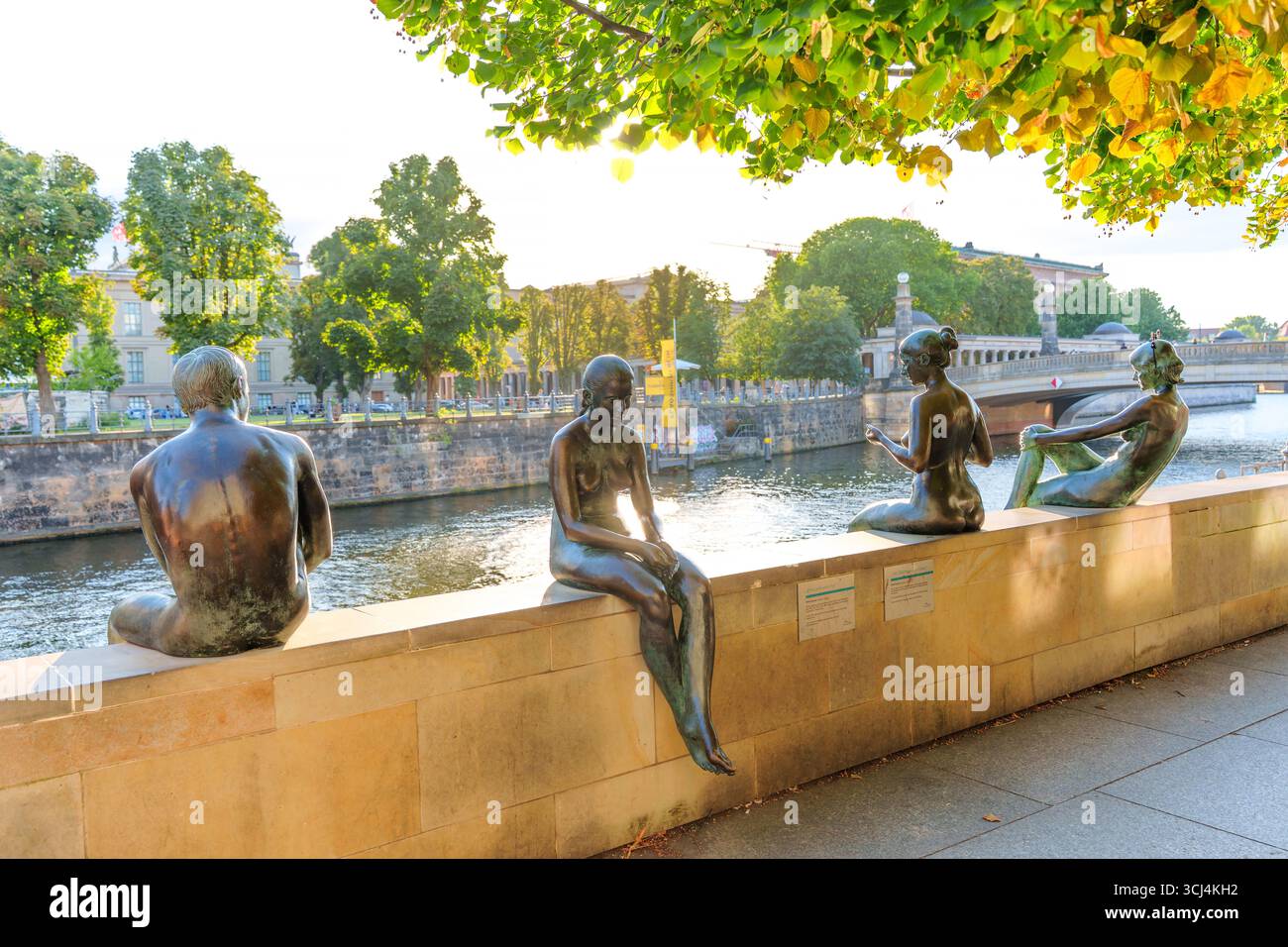 Berlino, Germania - 17 agosto 2025: Gruppo di statue in bronzo raffiguranti tre ragazze e un ragazzo seduti lungo il fiume Sprea, circondati da una vegetazione lussureggiante Foto Stock
