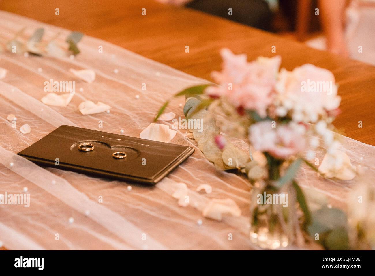 Le fedi nuziali sono elegantemente esposte su un vassoio scuro con petali floreali sparsi e delicati dettagli in tessuto. I fiori rosa tenui esaltano il romantico Foto Stock