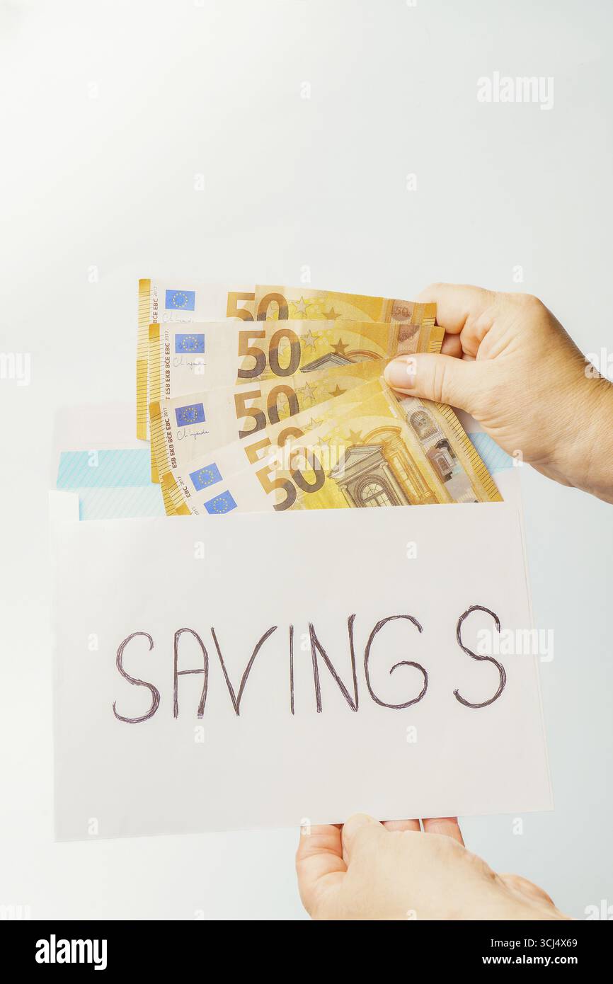Mani che inseriscono cinquanta banconote in euro in una busta bianca etichettata risparmio, illustrando la sicurezza finanziaria e la gestione del contante Foto Stock