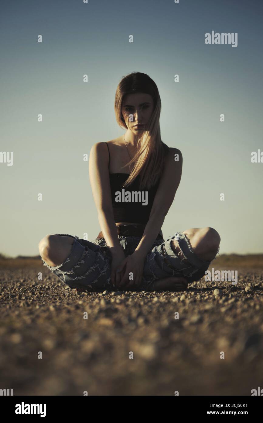 Una ragazza seria e triste, in jeans strappati, si siede su una strada deserta Foto Stock