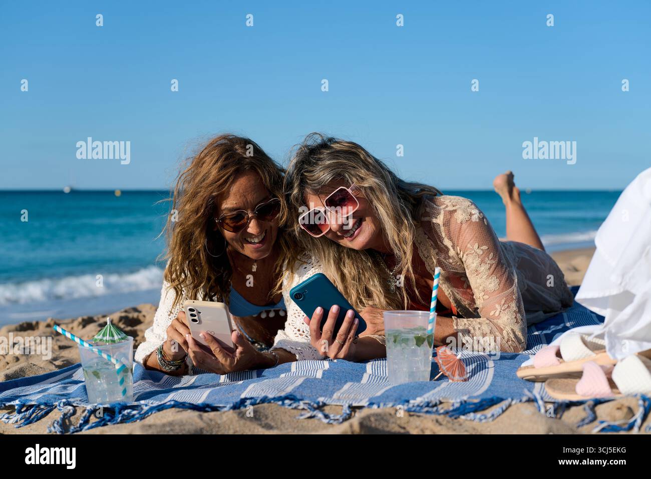 Due donne mature che utilizzano smartphone in spiaggia, si godono le vacanze estive Foto Stock