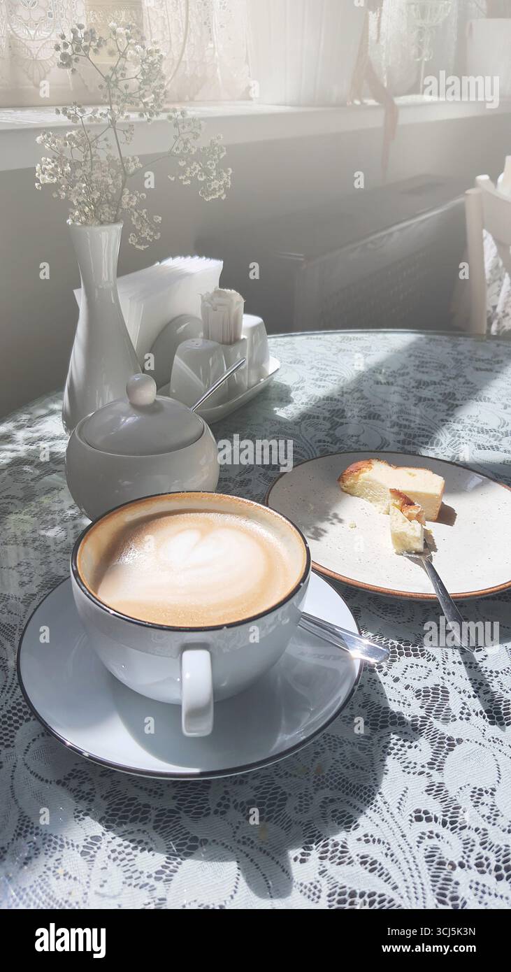 Goditi una tazza di caffè e una fetta di torta in un caffè caldo e invitante. La tovaglia in pizzo aggiunge fascino, mentre la luce del sole filtra attraverso la finestra, enha Foto Stock