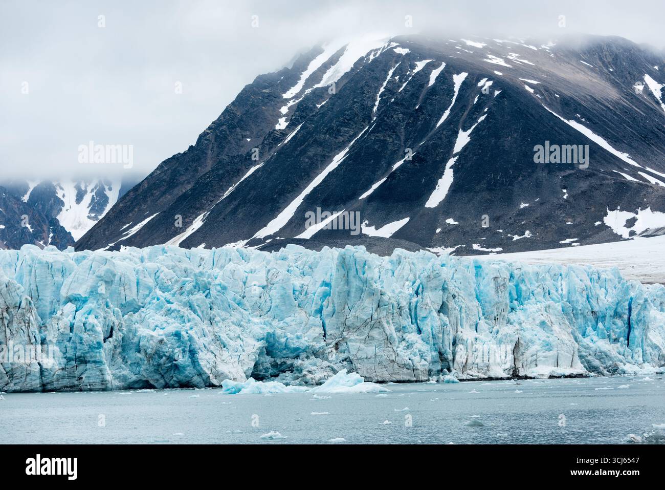 Il ghiacciaio Lilliehöökfjorden Spitsbergen Svalbard Norvegia // LILLIEHÖÖKFJORDEN, Svalbard, Norvegia — Una massiccia parete glaciale, caratterizzata da ghiaccio blu profondo e crepacci spettacolari, fronteggia una catena montuosa rocciosa scura parzialmente ricoperta di neve e avvolta dalla nebbia. I vitelli del ghiacciaio si iceberg nelle frigide acque artiche, una vista comune in questo remoto arcipelago. Lilliehöökfjorden si trova sulla costa nord-occidentale di Spitsbergen, l'isola più grande dell'arcipelago delle Svalbard. Le Svalbard sono un territorio norvegese situato nell'Oceano Artico, noto per i suoi ghiacciai, gli orsi polari e il paesaggio ghiacciato Foto Stock