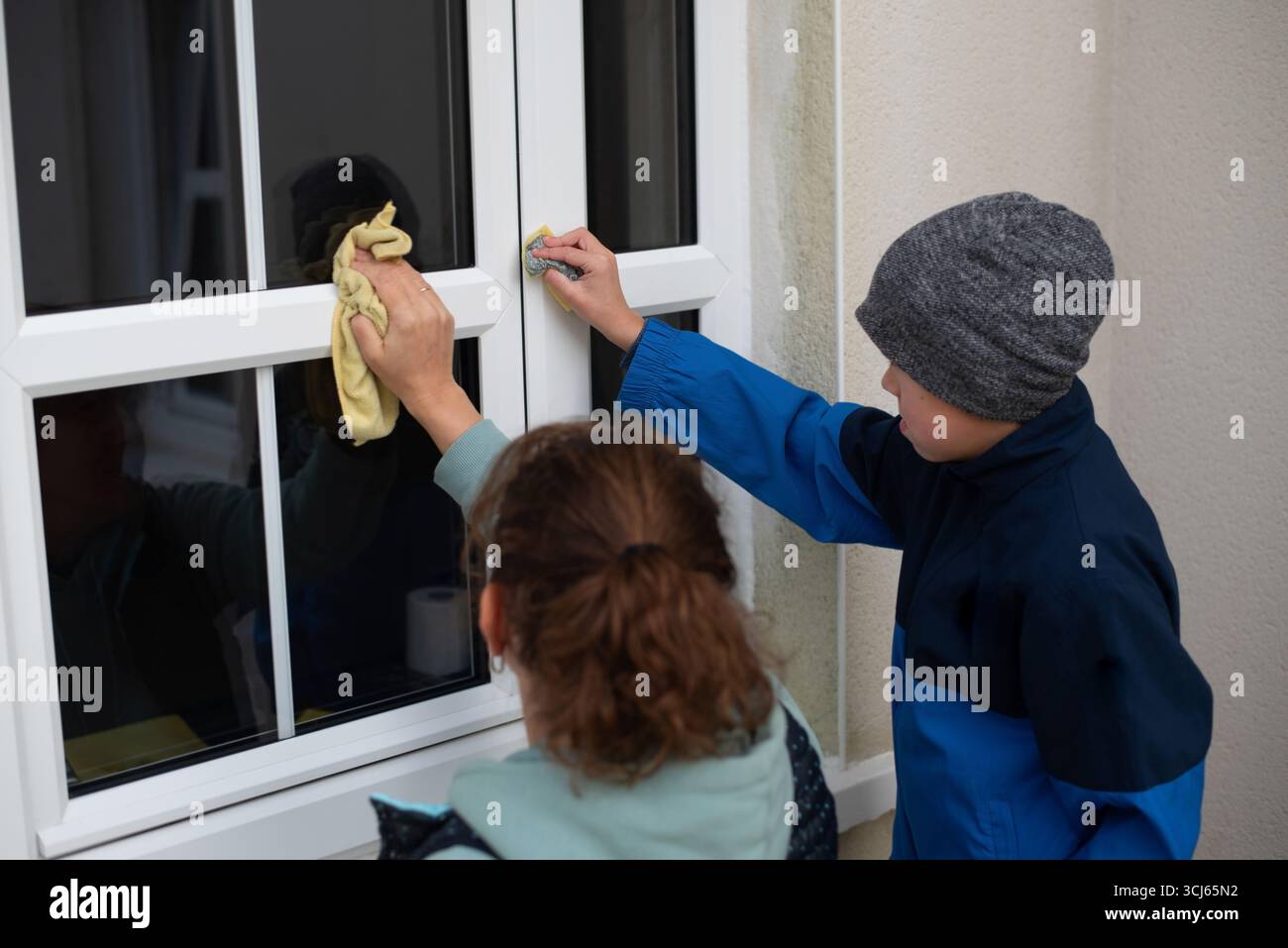 Due bambini puliscono una finestra all'aperto con un panno e una spugna. Stanno lavorando insieme per un'attività di pulizia, mostrando cooperazione e risposte Foto Stock