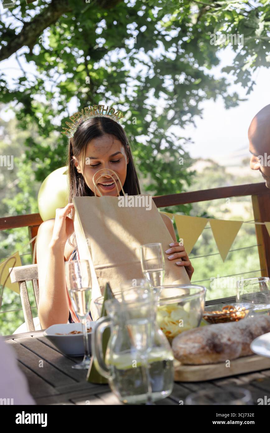 Donna che apre una borsa regalo alla festa all'aperto, sorridente e circondata da amici Foto Stock