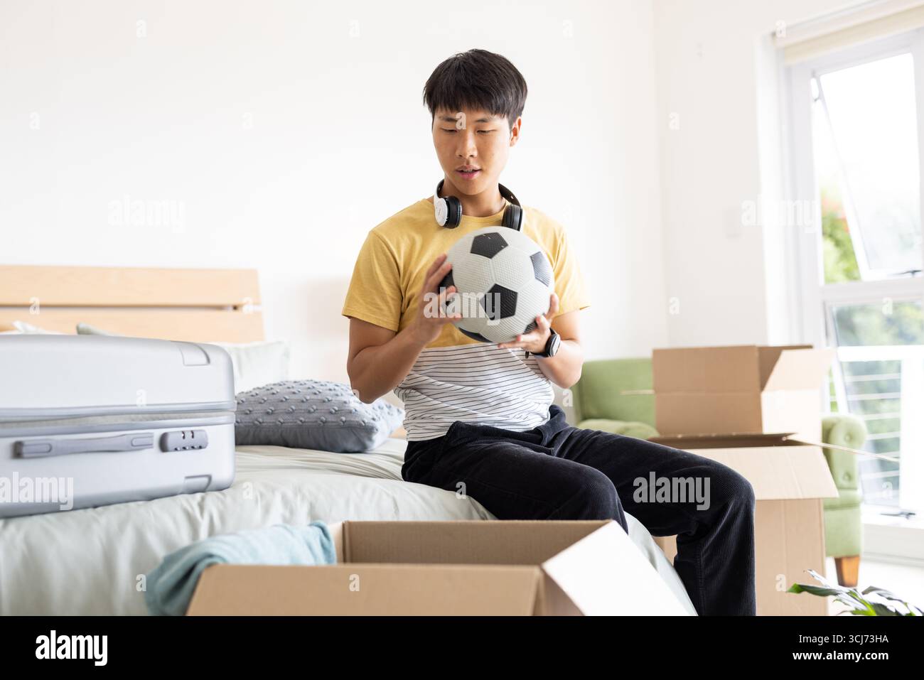 Disimballaggio delle scatole in camera da letto, giovane che tiene la palla da calcio con le cuffie Foto Stock