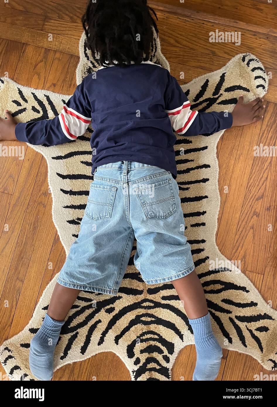 Primo piano di un ragazzino che gioca su un tappeto di tessuto umoristico che sembra una pelle di tigre su un pavimento di legno duro, 2025, NYC, USA Foto Stock