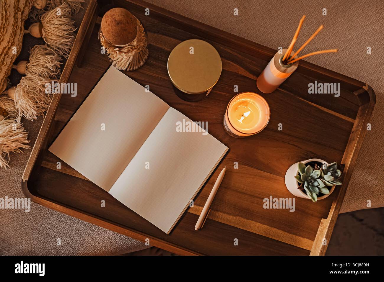 Accogliente area di lavoro con quaderno aperto per il diario su un vassoio di legno circondato da candele profumate e una piccola pianta. Foto Stock