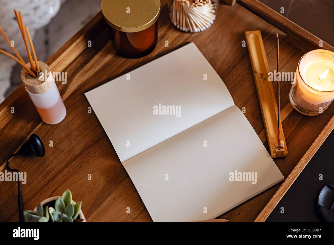Accogliente area di lavoro con quaderno aperto per il diario su un vassoio di legno circondato da candele profumate, incenso e una piccola pianta. Foto Stock