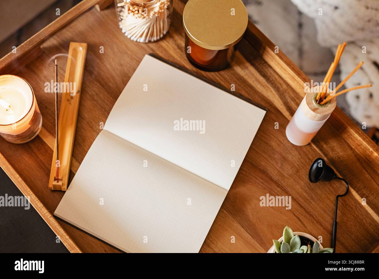 Spazio di lavoro per il diario o la meditazione con un quaderno aperto su un vassoio di legno circondato da candele profumate, incenso e una piccola pianta Foto Stock