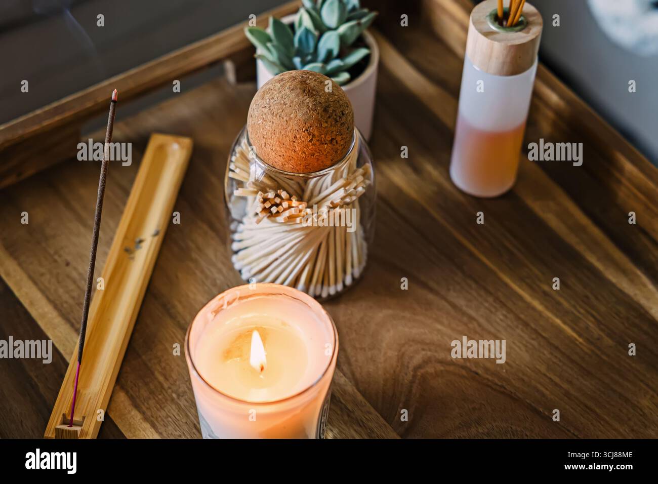 Una scena calda e invitante con una candela illuminata, un bastone di incenso e un barattolo di fiammiferi su un vassoio di legno, rituale di meditazione mattutino Foto Stock