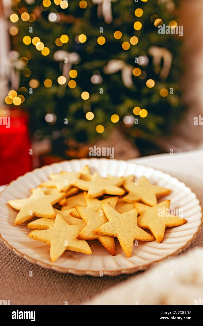 Un piatto di biscotti a forma di stella è esposto di fronte a un albero di Natale splendidamente illuminato, creando un'atmosfera di vacanza calda e festosa. Foto Stock