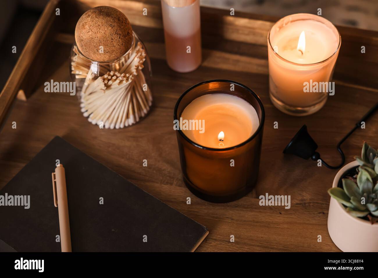 Una scena calda e invitante con candele accese, blocco note per il diario, un vaso di fiammiferi, un quaderno con penna e una piccola pianta su una scrivania di legno. Foto Stock