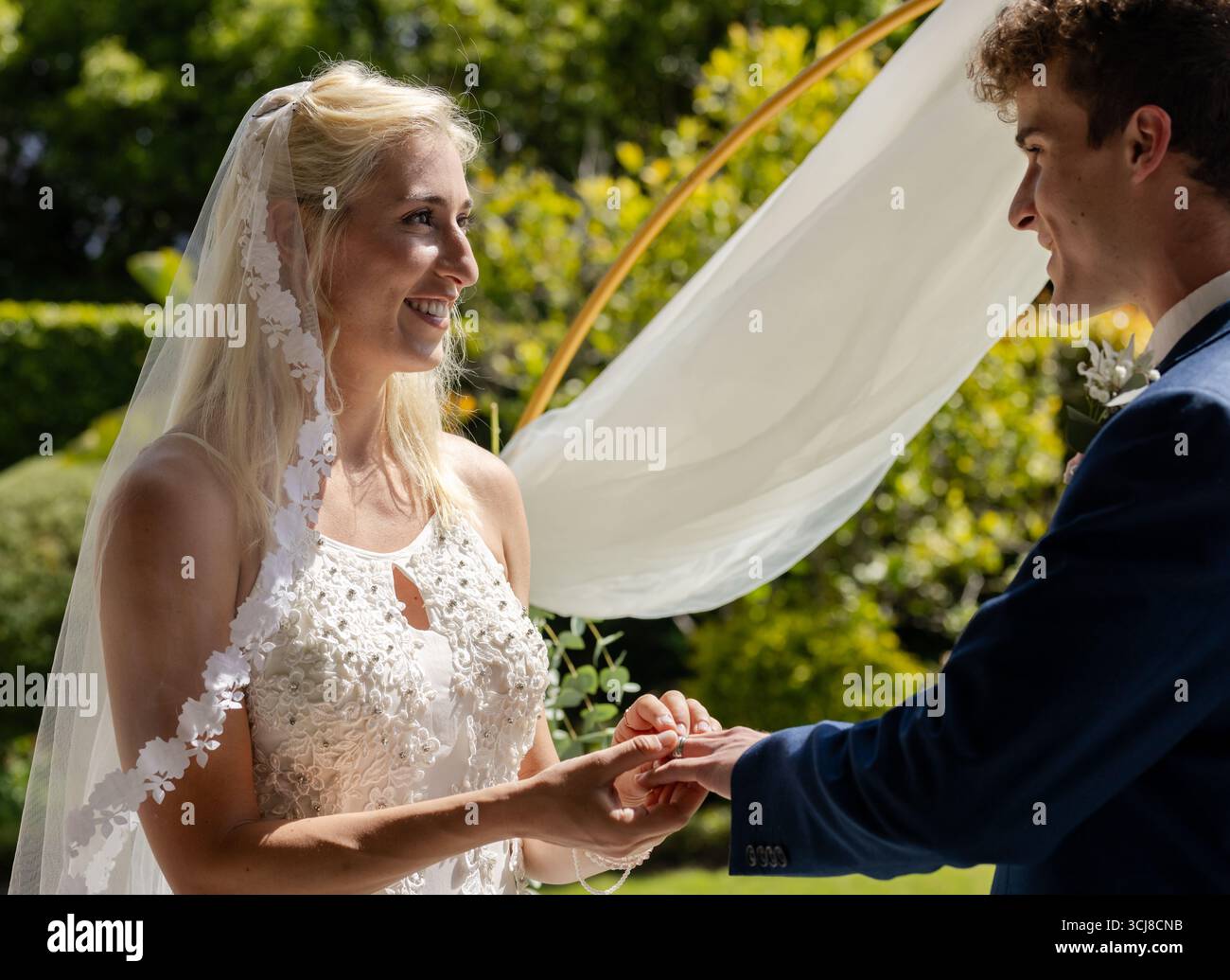 La sposa si scambia anelli con lo sposo all'aperto, entrambi sorridenti alla luce del sole Foto Stock