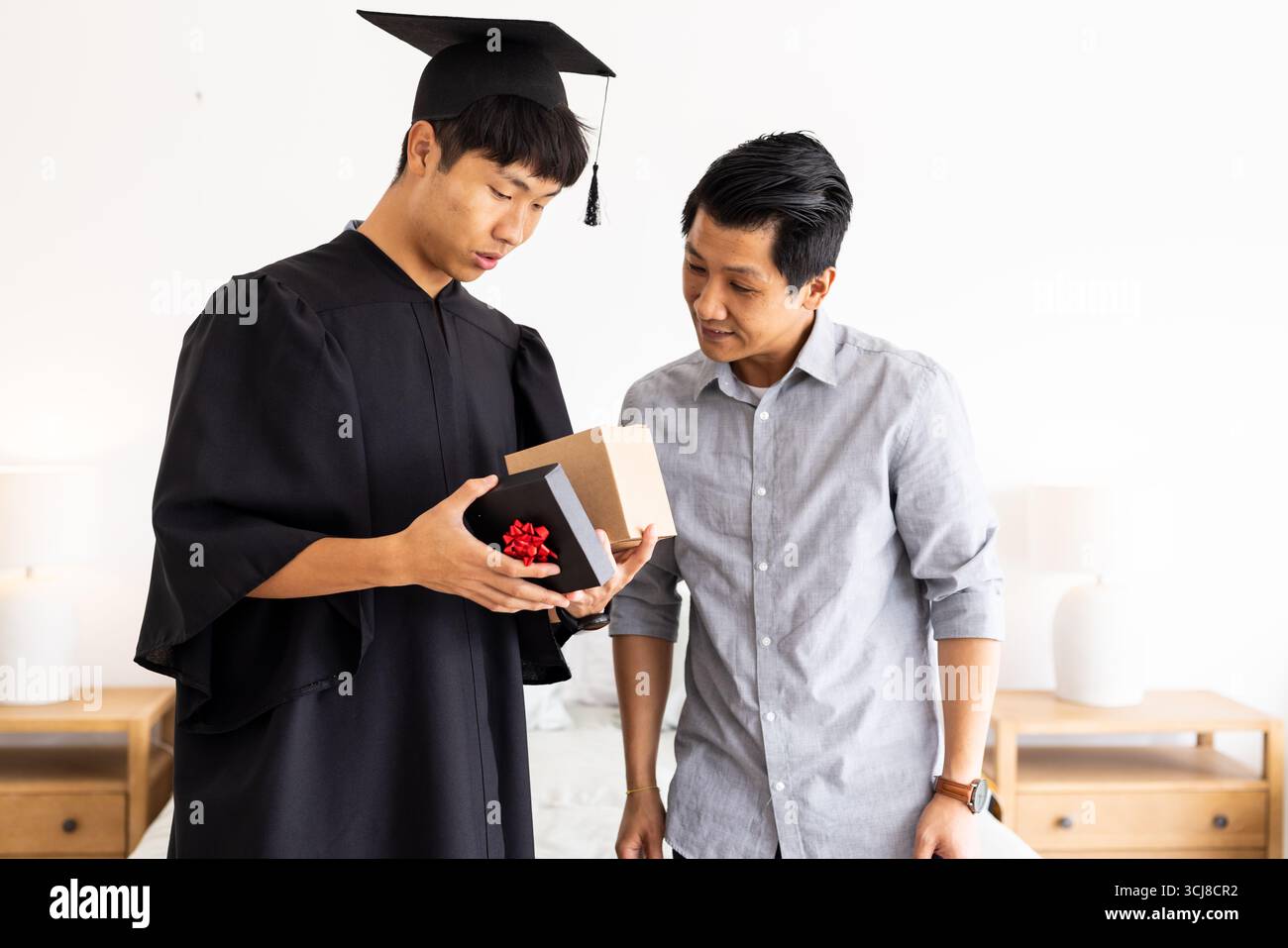 Laureati in berretto e abito con il padre a casa, sentendosi sorpreso Foto Stock