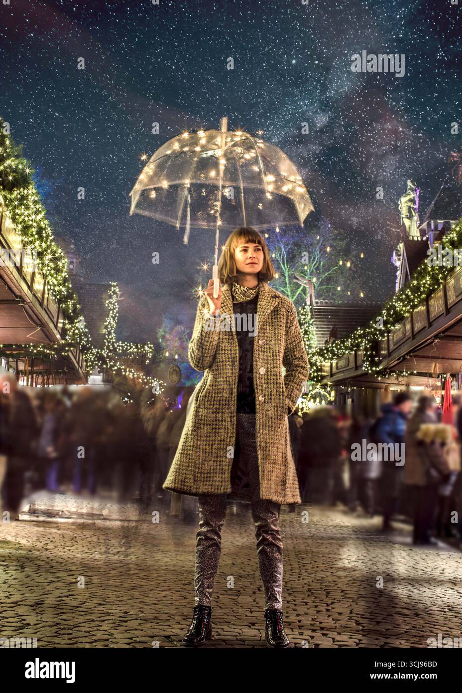 Bella donna alla moda in piedi nel bel mezzo del mercatino di Natale, mentre tiene in mano un ombrello trasparente con luci da favola. Fotografato di notte. Foto Stock