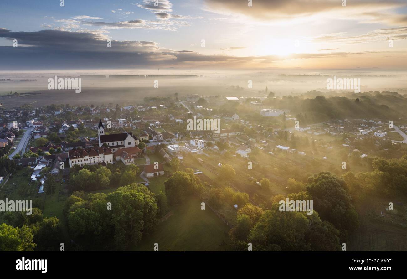 Foto droni dell'alba nebbiosa sulla foresta, sul lago e sul villaggio - luce del sole dorata con raggi e ombre. Foto Stock