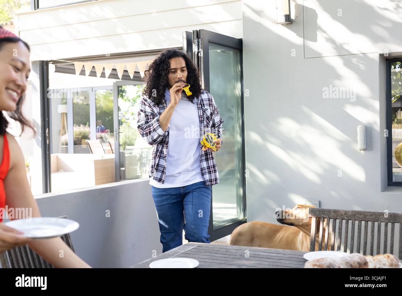 Uomo che si gode uno spuntino all'aperto mentre donna prepara un tavolo per festeggiare con il cane nelle vicinanze Foto Stock