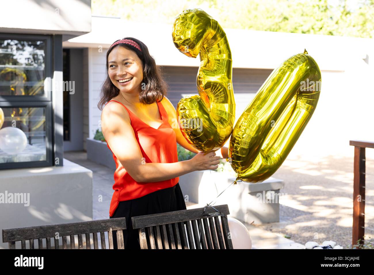 Donna asiatica con palloncini d'oro che festeggiano il compleanno all'aperto, sorridendo gioiosamente Foto Stock