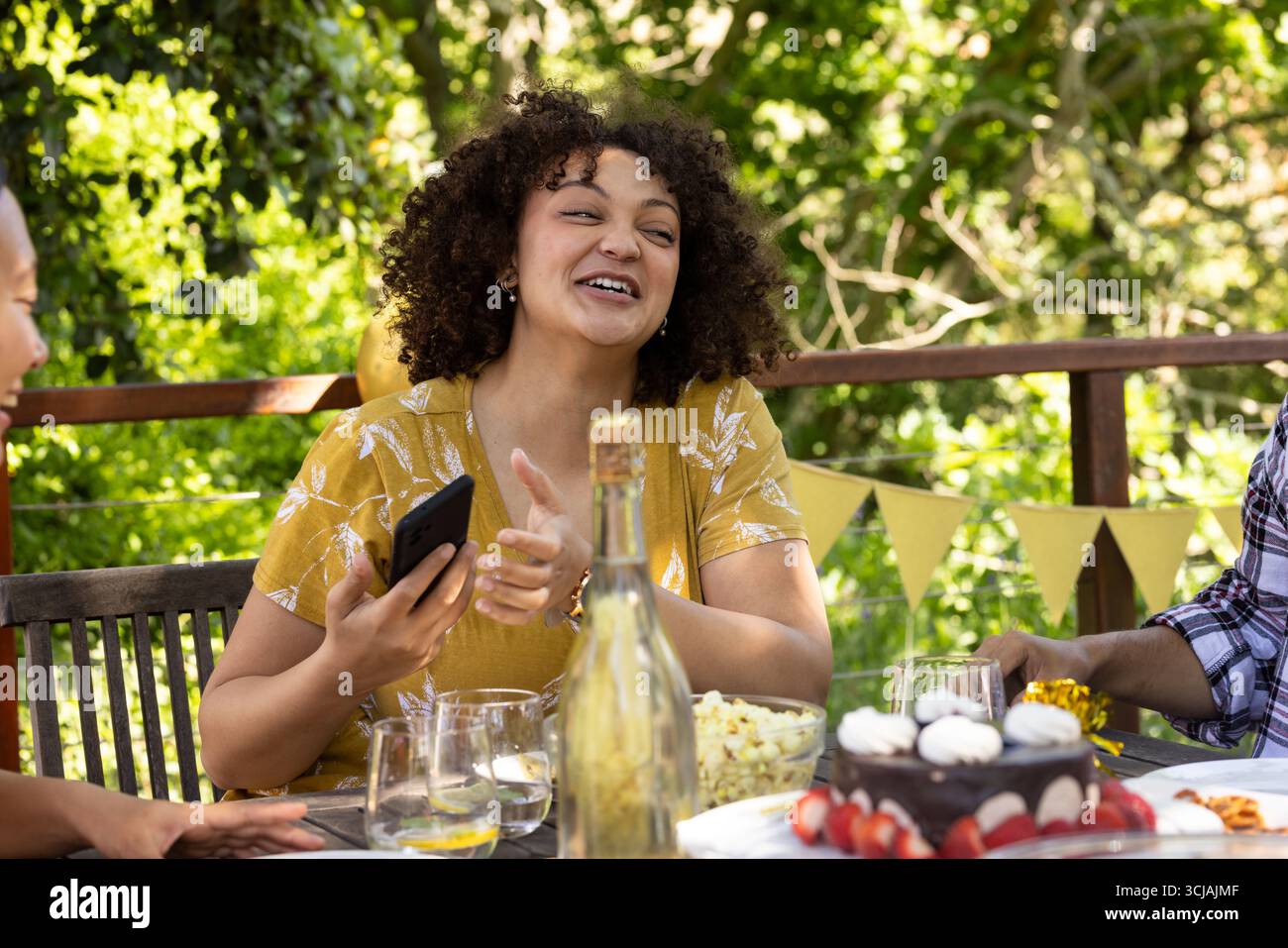 Donna ridendo che tiene in mano lo smartphone alla festa all'aperto con gli amici e la torta Foto Stock