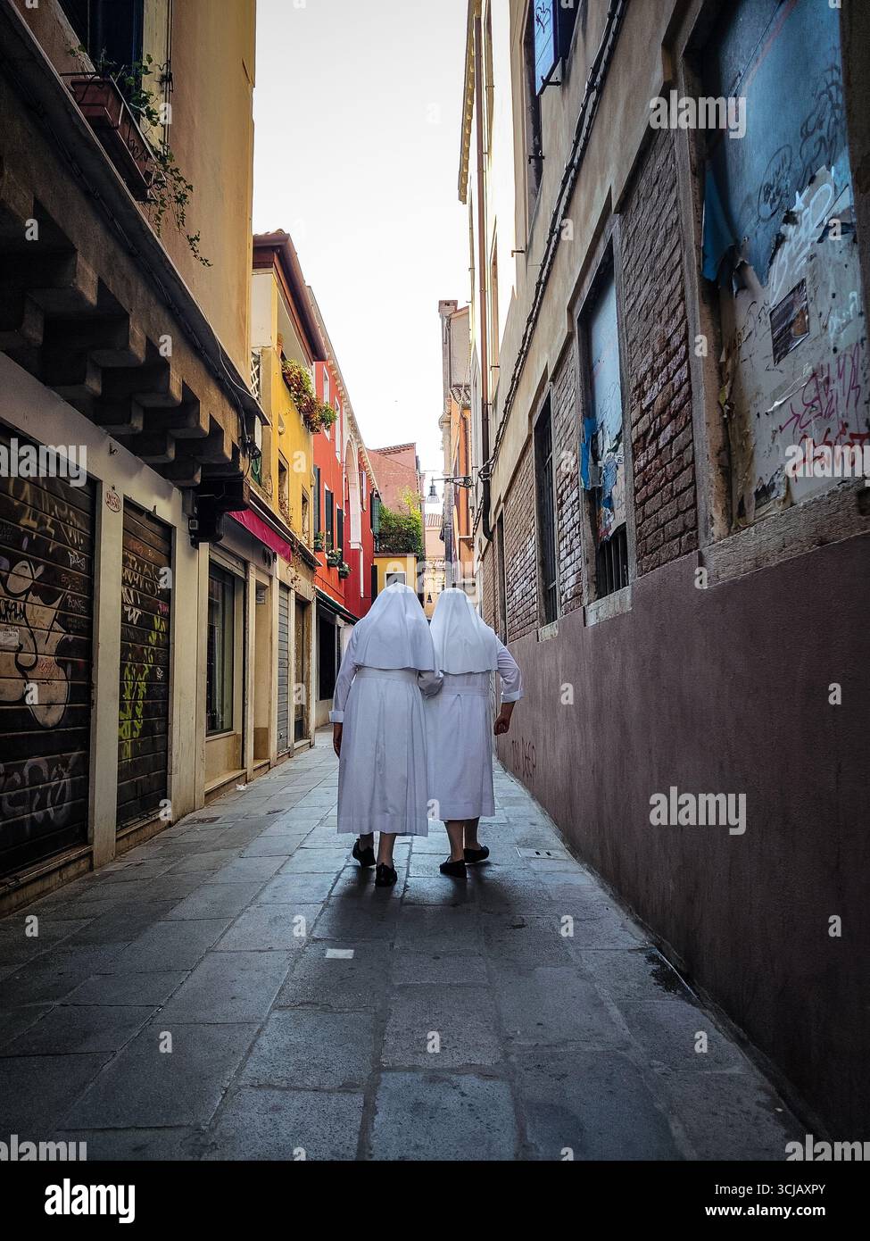 Due suore in abiti bianchi camminano lungo uno stretto e storico vicolo di Venezia, Italia. L'immagine cattura un momento sereno e senza tempo, con le figure stan Foto Stock