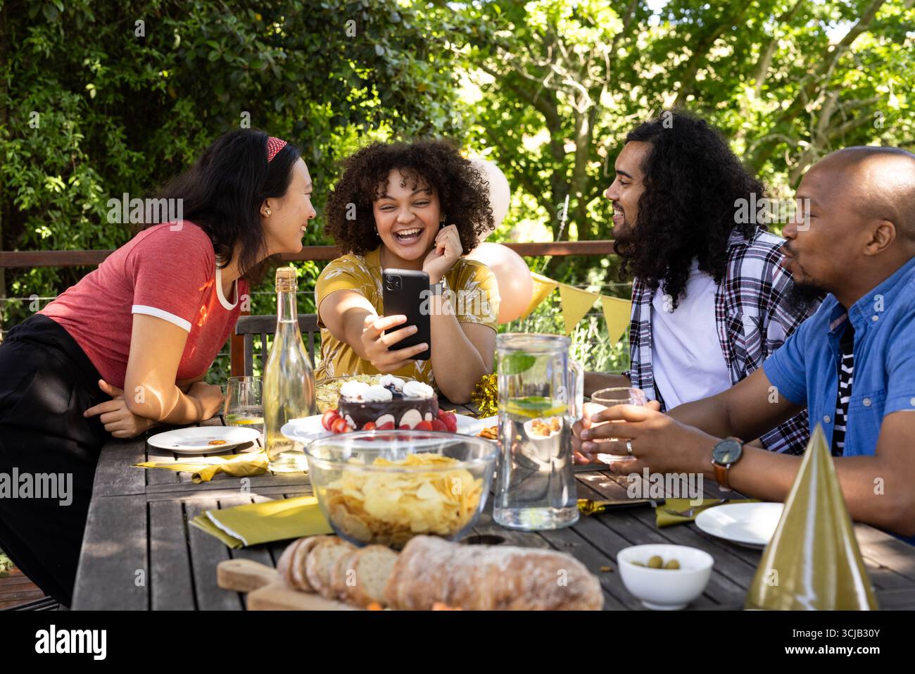 Festeggia all'aperto, gli amici ridono e scattano selfie al tavolo festivo con spuntini Foto Stock