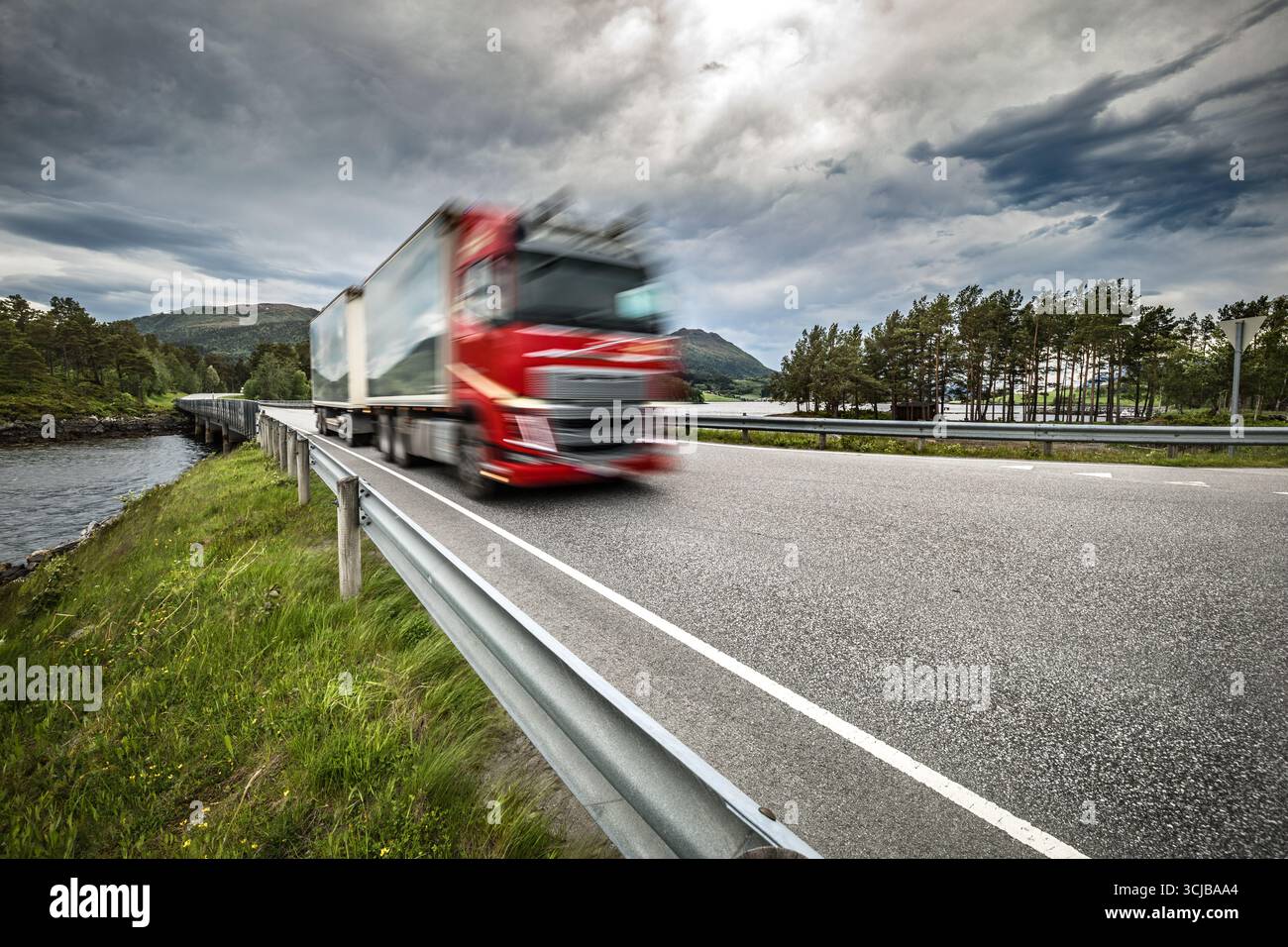Il semi-camion rosso sta percorrendo una strada con un cielo nuvoloso sullo sfondo. Il carrello si muove rapidamente e la strada è costeggiata da alberi. La scena è fas Foto Stock
