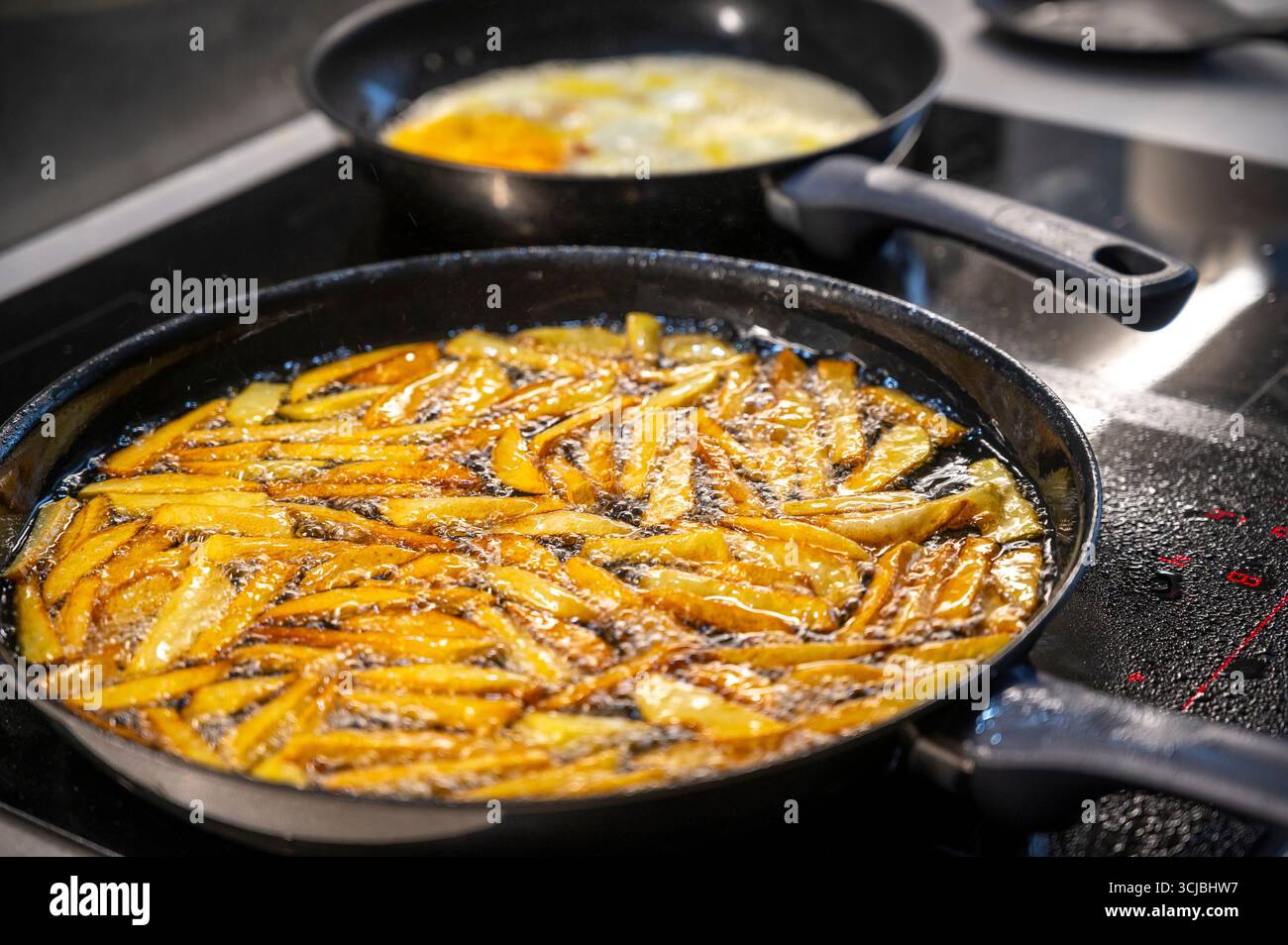 Le patatine dorate spolverano l'olio caldo all'interno di una padella nera su un piano cottura. La scena della cucina cattura il momento della cottura con le bollicine che formano un Foto Stock