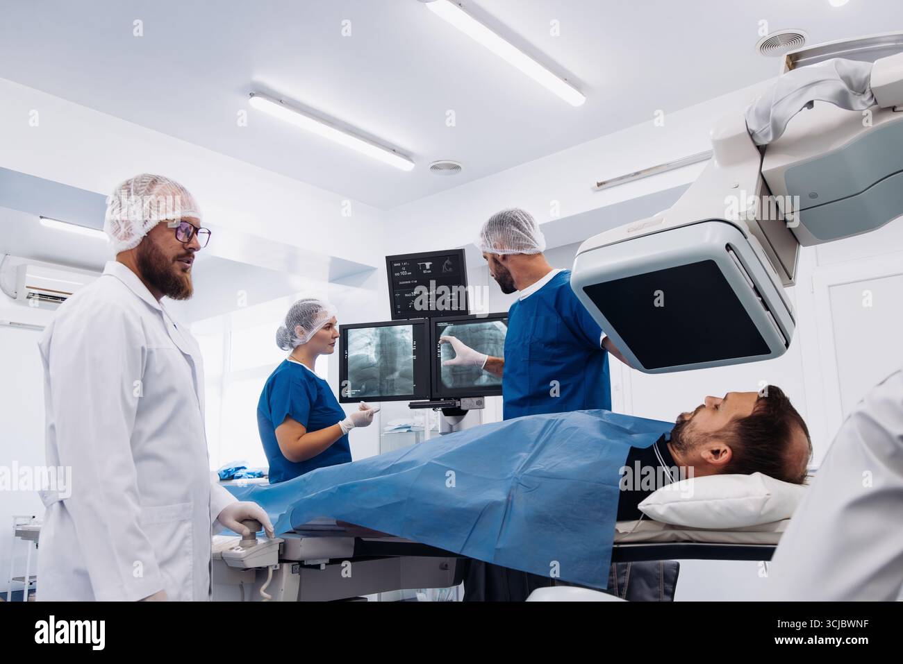 I professionisti del settore medicale in scrub e camici da laboratorio si concentrano sull'analisi delle immagini angiografiche visualizzate sugli schermi durante la cura di un paziente su un medico Foto Stock
