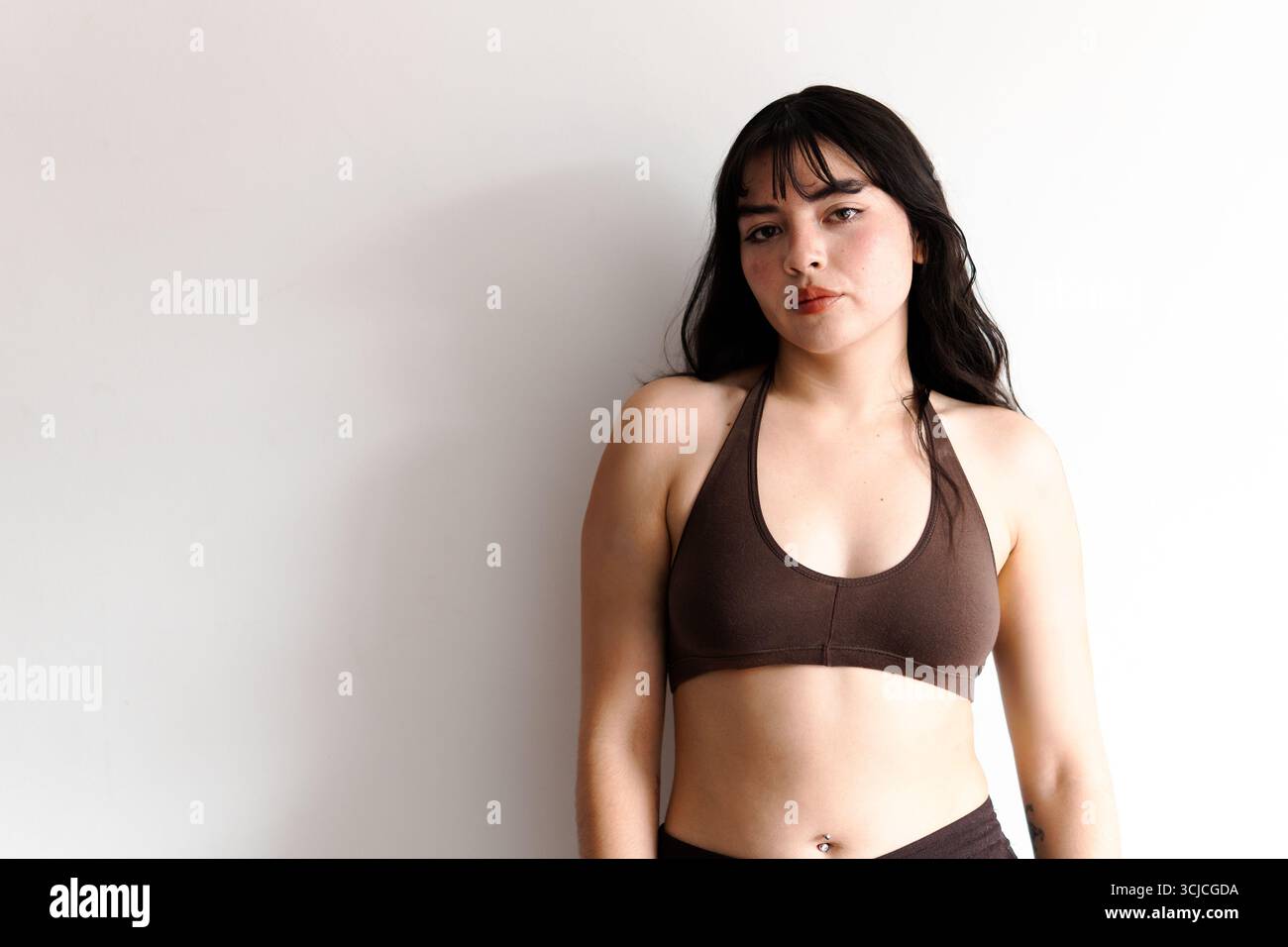 Una donna sicura di sé con lunghi capelli scuri si staglia contro una parete bianca, guardando la macchina fotografica e indossando una parte superiore marrone. La sua posa rilassata trasmette forza e Foto Stock
