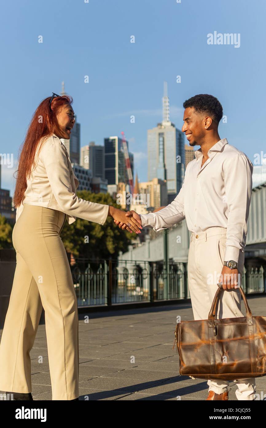 Un uomo e una donna, vestiti con abiti business casual, stringono la mano all'aperto di fronte allo skyline di Melbourne, esemplificando gli affari multiculturali Foto Stock