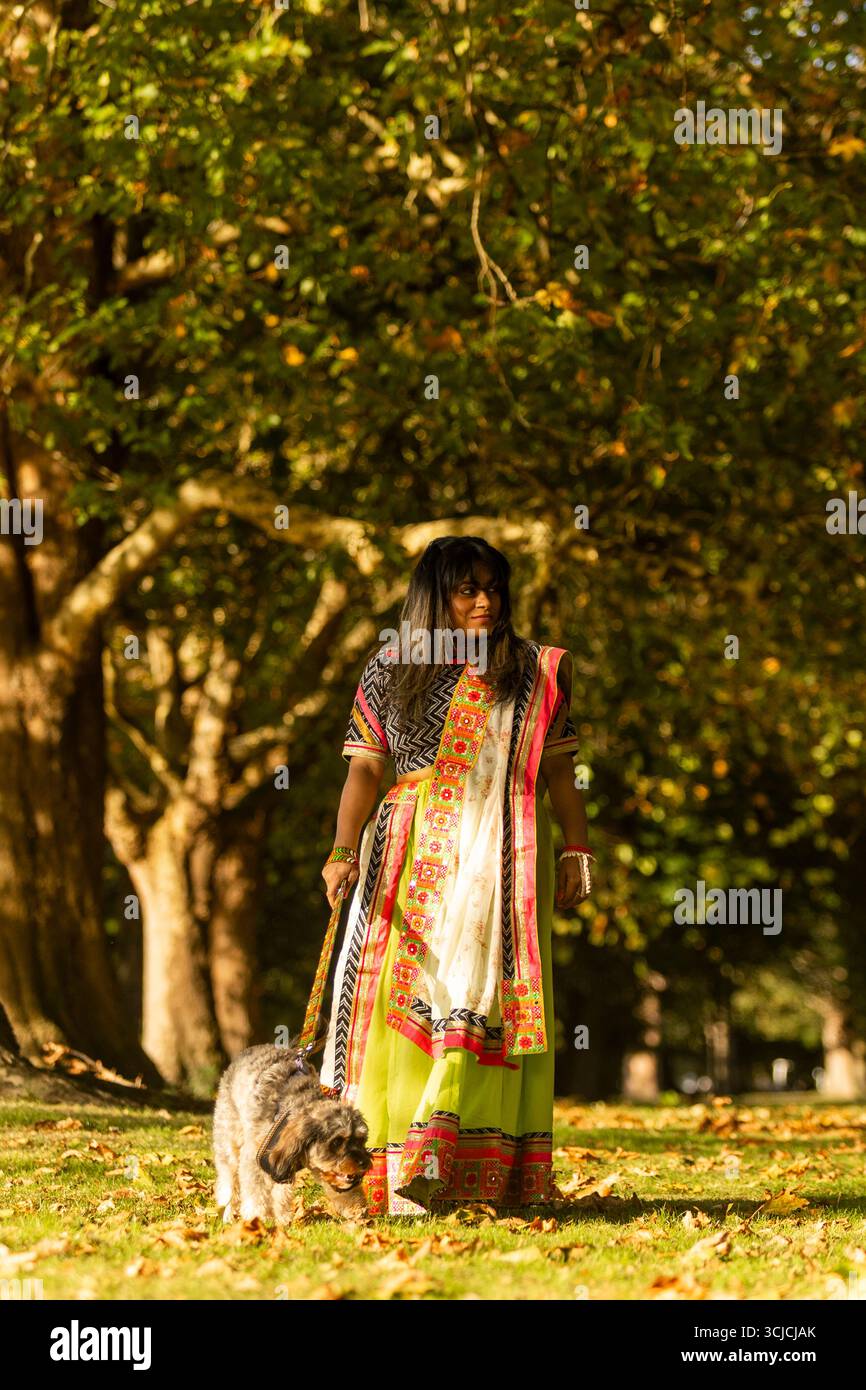 Donna indiana in un colorato sari che porta il suo cane in un parco autunnale. Il sole filtra attraverso gli alberi, emettendo un caldo bagliore, creando un'immagine serena Foto Stock