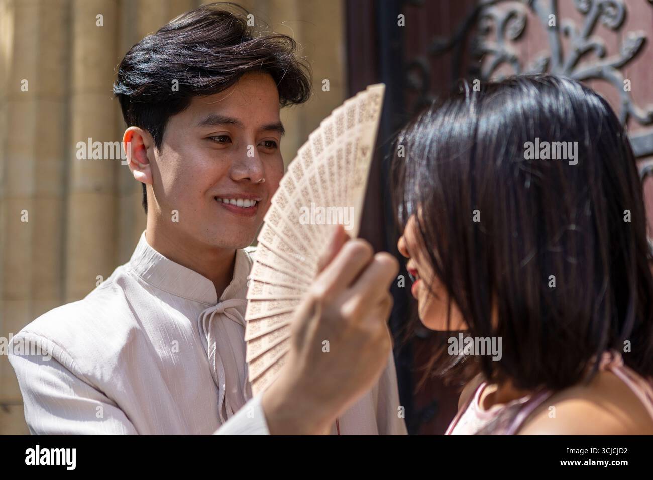 Una coppia vietnamita condivide un momento tenero a Melbourne, con l'uomo che tiene giocosamente un fan in faccia alla donna. Indossano abiti tradizionali Foto Stock