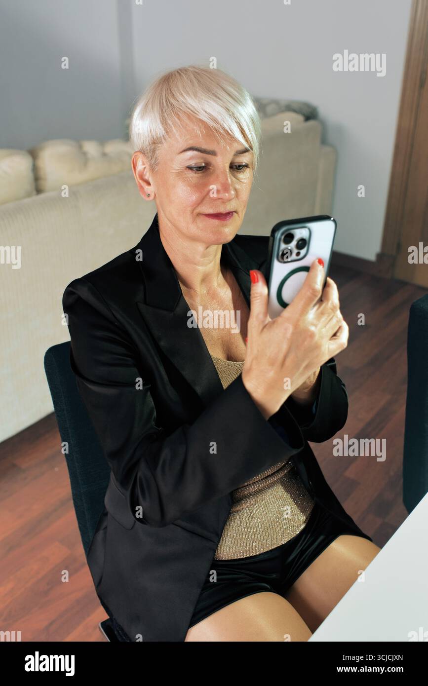 Una donna con i capelli corti concentrata sul suo smartphone mentre lavora online in un ufficio domestico. È seduta a un tavolo, vestita con un abbigliamento casual elegante, Re Foto Stock