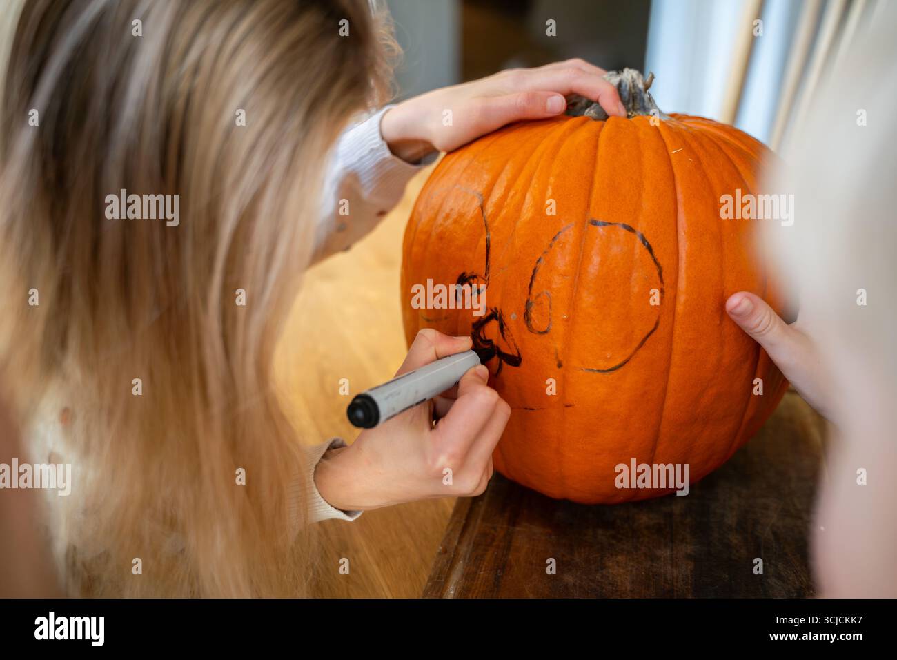 Una giovane donna disegna con cura un disegno di Halloween su una grande zucca arancione usando un pennarello nero, preparandolo per l'intaglio. La scena cattura la f Foto Stock