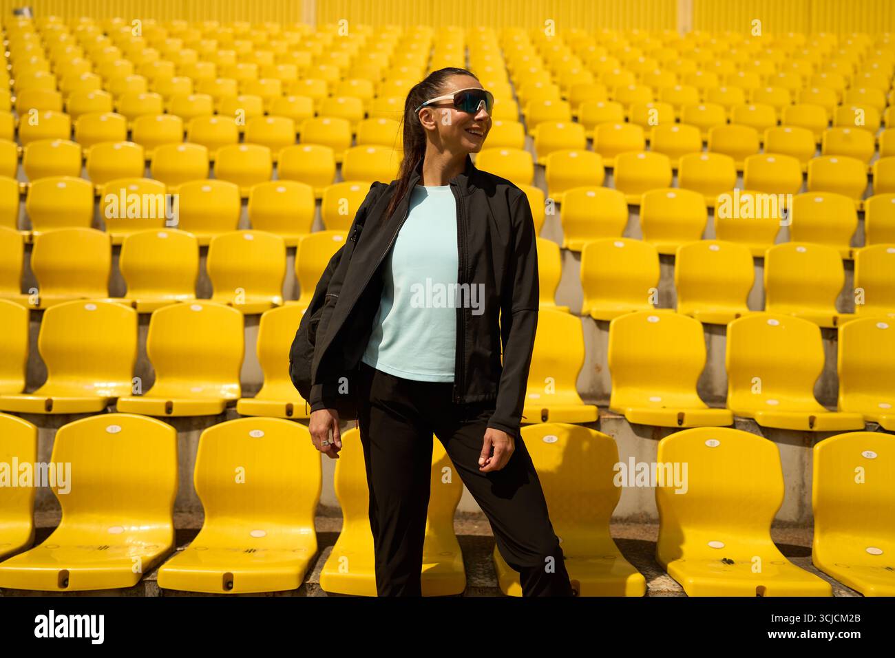 Un'atleta sicura di sé sorride in uno stadio soleggiato, circondato da vibranti sedili gialli. Incarna uno stile di vita attivo, irradiando positivi Foto Stock