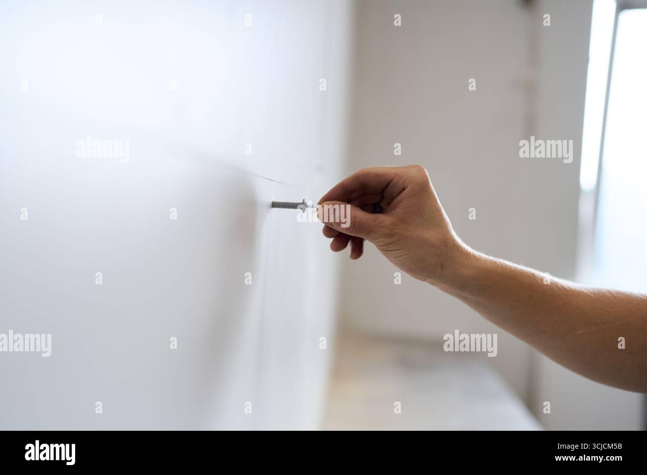 La mano di un uomo fissa con cura una vite in un muro bianco, indicando le fasi iniziali di un progetto di ristrutturazione. Rappresentazione perfetta di precisione e. Foto Stock