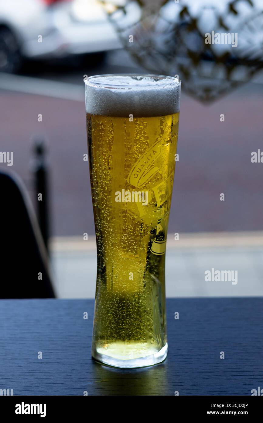 primo piano di un alto bicchiere di birra sudato con una testa spumeggiante e bolle in lievitazione, seduti su un tavolo scuro. Foto Stock