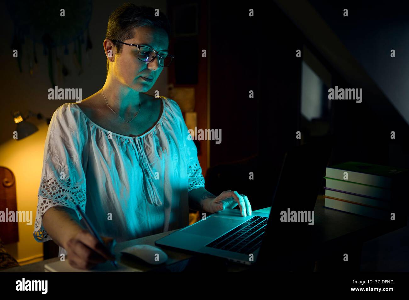 Una donna di mezza età si siede alla scrivania di notte, lavorando sul proprio notebook in un ambiente di ufficio domestico poco illuminato. La luce calda della lampada crea un bancomat mirato Foto Stock