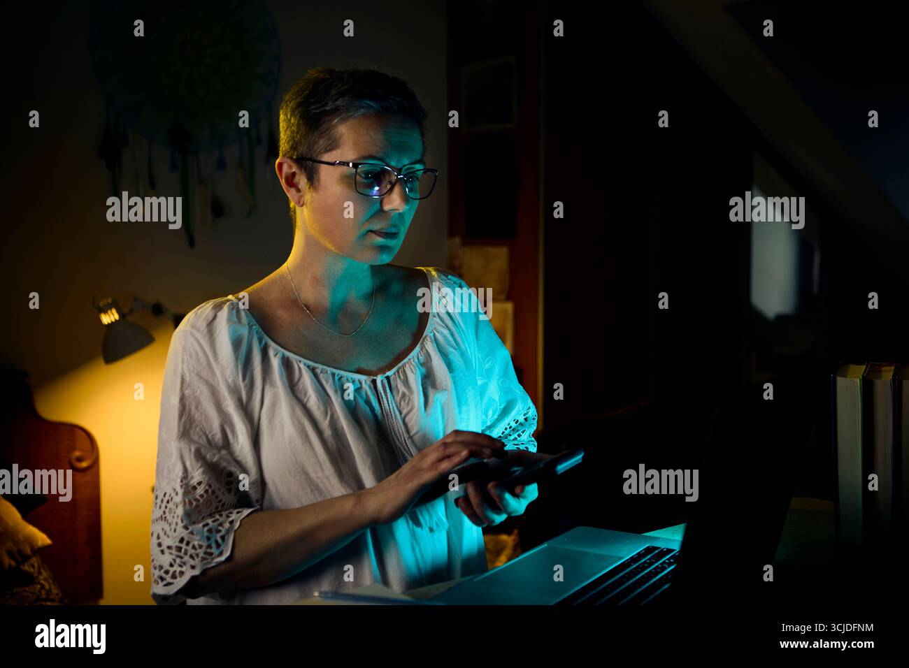 Una donna di mezza età con una camicetta bianca si indossa sul suo laptop mentre lavora da casa la sera. La calda lampada da scrivania mette in risalto la sua attenzione, rappresentativa Foto Stock
