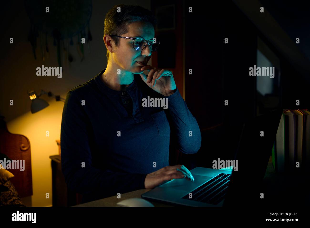 Una donna di mezza età lavora in remoto sul suo notebook tenendo il telefono e multitasking in un ufficio buio. L'immagine rappresenta un'opera moderna di ho Foto Stock