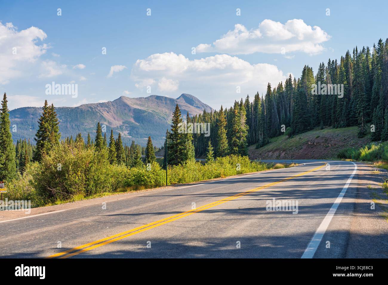 Una strada panoramica di montagna si snoda con grazia attraverso una lussureggiante e vibrante foresta verde, che conduce verso vette maestose sotto un cielo blu brillante punteggiato di così Foto Stock