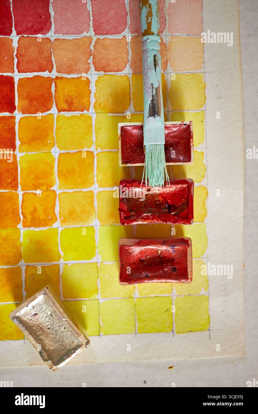 Primo piano di campioni di vernice ad acquerello in tonalità calde con teglie e pennello rossi. Composizione artistica vibrante, perfetta per progetti creativi e design Foto Stock