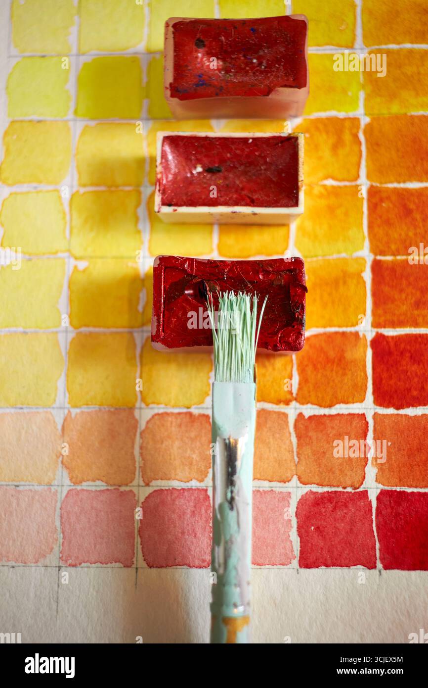 Primo piano di campioni di vernice ad acquerello in tonalità calde con teglie e pennello rossi. Composizione artistica vibrante, perfetta per progetti creativi e design Foto Stock