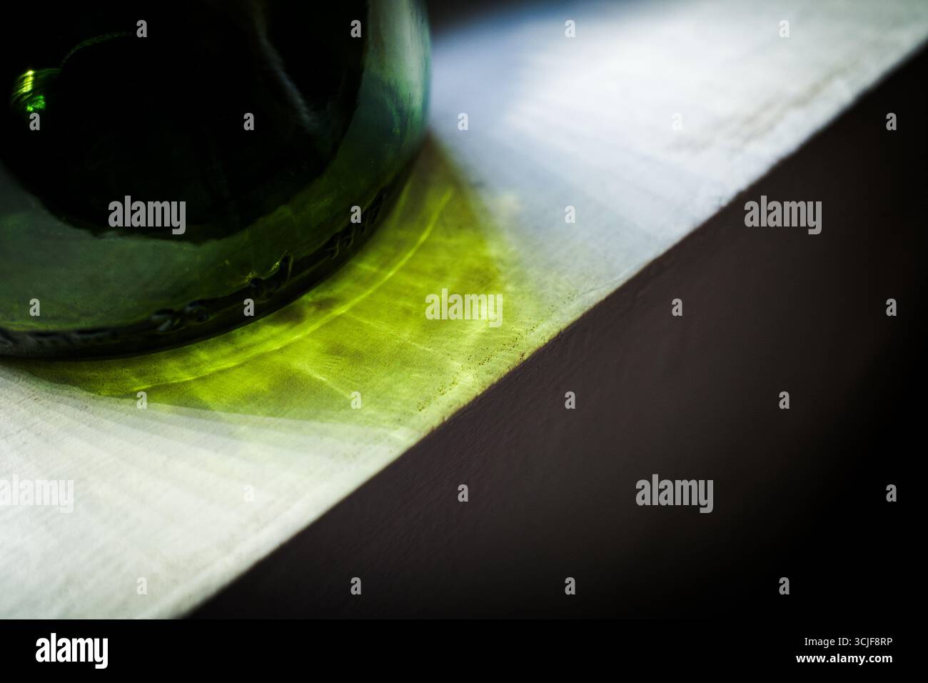 Primo piano della luce del sole che passa attraverso una bottiglia di vetro verde, creando riflessi gialli-verdi astratti e ombre su una superficie bianca. Foto Stock