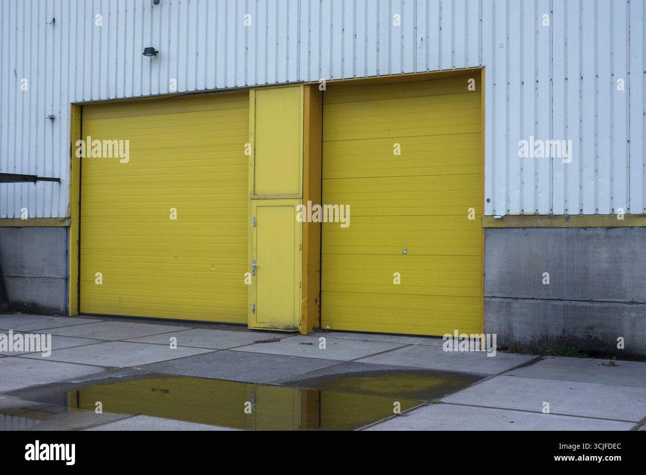 Due grandi banchine di carico di colore giallo brillante su un edificio industriale in metallo ondulato si riflettono in una pozzanghera sulla pavimentazione in cemento Foto Stock