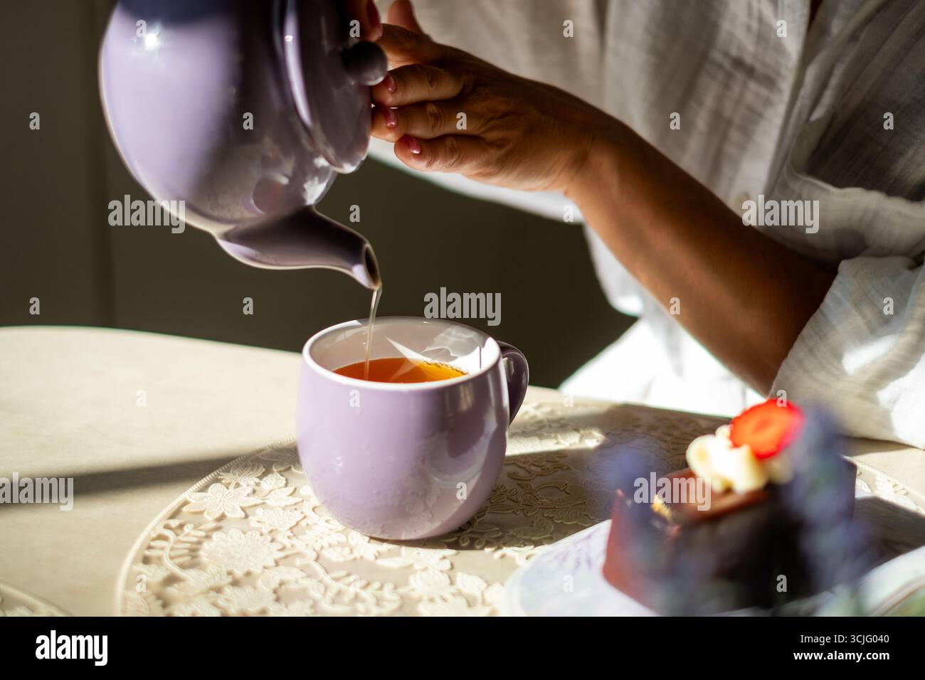 La figura femminile versa con grazia il tè in una tazza viola, con pasticcini nelle vicinanze su una tovaglia di pizzo, creando un'atmosfera serena di gioia ed eleganza Foto Stock