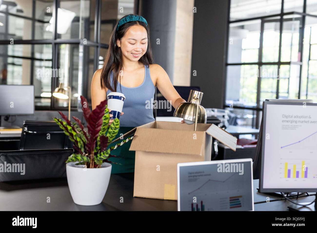 Donna asiatica che disimballa la scatola in un ufficio moderno, tiene una tazza di caffè e sorride Foto Stock