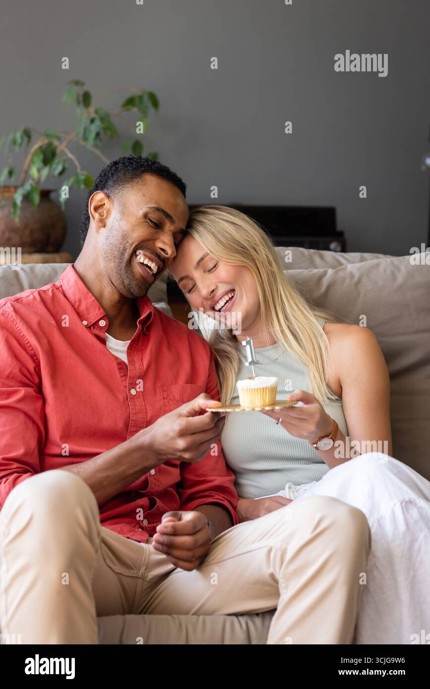 Coppia sul divano che condivide il cupcake, ridendo e godendosi un'accogliente festa a casa Foto Stock